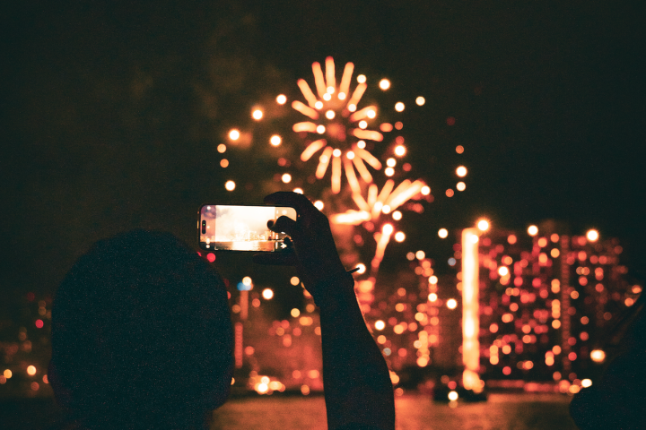 Silhouette of person photographing fireworks over city skyline at night.