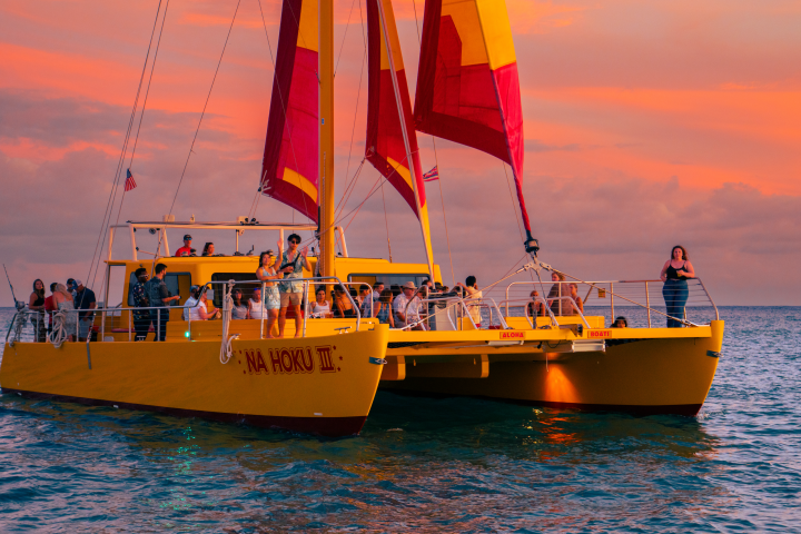 People on a yellow catamaran with red sails at sunset on the ocean.